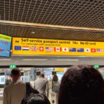 Photograph of travelers queuing at self-service passport control gates beneath overhead signs in an airport terminal.