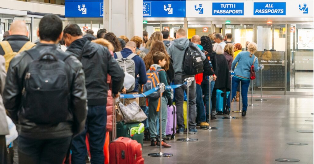 Travellers line up with luggage at an airport passport control area under signs reading all passports.