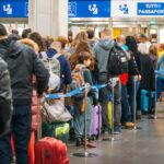 Travellers line up with luggage at an airport passport control area under signs reading all passports.