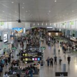 Busy airport terminal with travelers walking, waiting, and lining up beneath overhead directional signs.