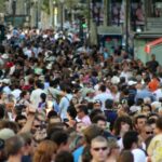 Crowded pedestrian street filled with people, kiosks, and trees, suggesting a busy city center.