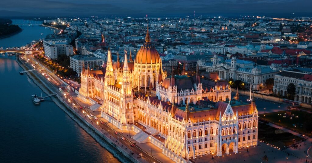 Illuminated historic parliament building along a river at night, with city lights and bridges visible in the background.