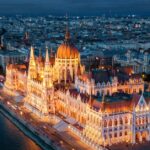 Illuminated historic parliament building along a river at night, with city lights and bridges visible in the background.