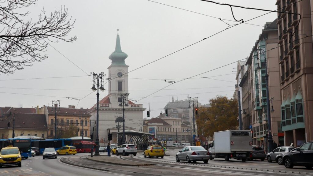 Busy city intersection with cars and trams, featuring a church tower and historic buildings on an overcast day.