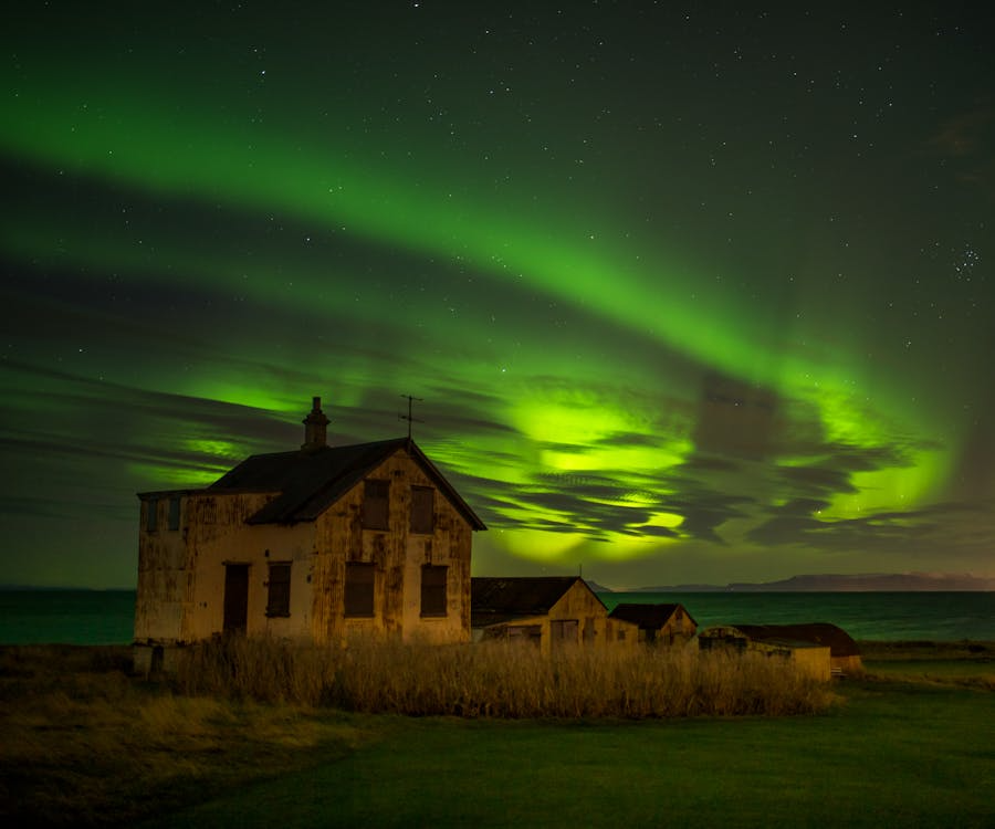 An abandoned house near the coast illuminated by green northern lights swirling across a star-filled night sky.
