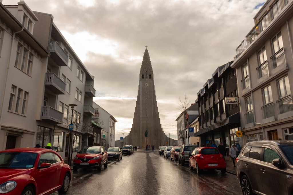 Hallgrímskirkja church rises at the end of a wet city street lined with parked cars and modern buildings under a cloudy sky.