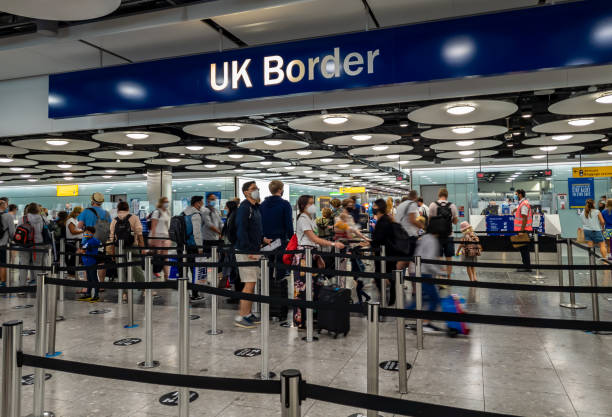 Travelers queue at a UK Border control area inside an airport, with overhead signage and security barriers visible.