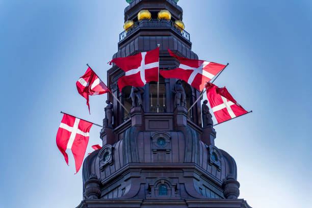 Multiple Danish flags fly around the top of a historic tower against a clear blue sky.