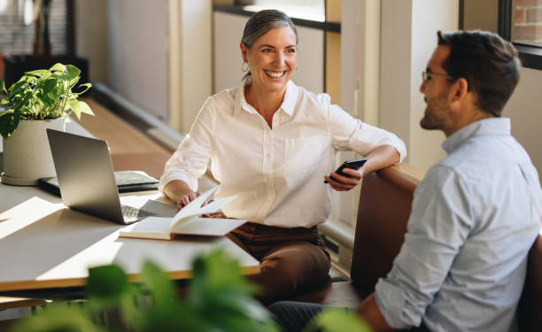 Two coworkers sit at a table discussing work, with a laptop, notebook, and phone in a bright office setting.