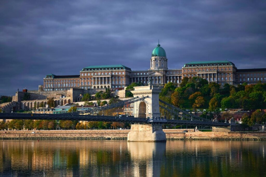 Historic castle complex on a hill overlooking a river and suspension bridge under a cloudy sky.