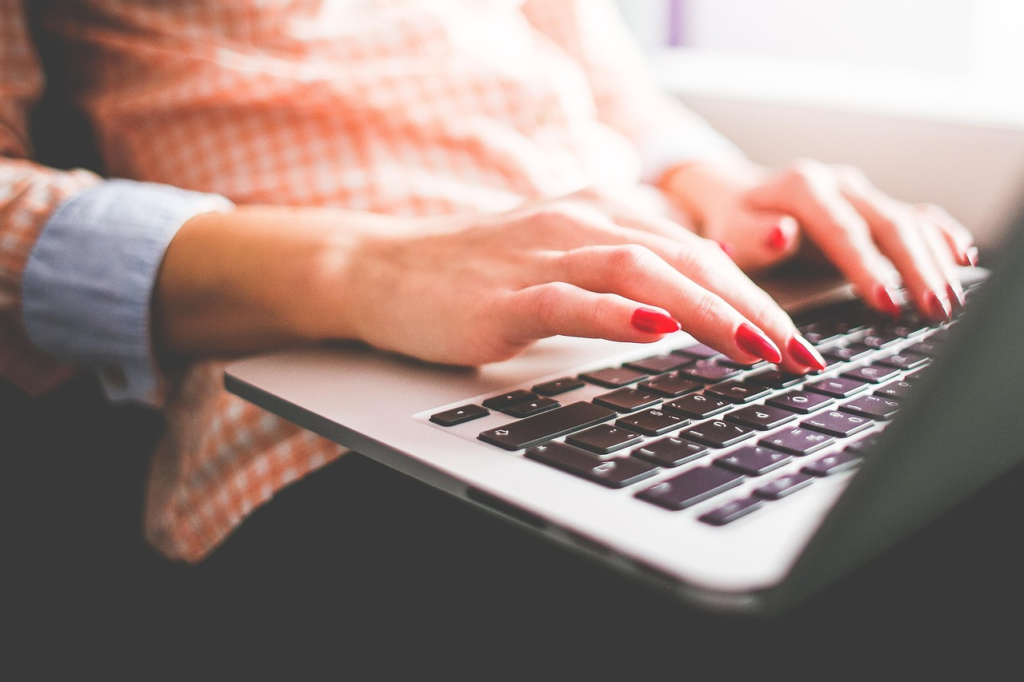 Close-up photo of hands with red nail polish typing on a laptop keyboard in a softly lit indoor setting.