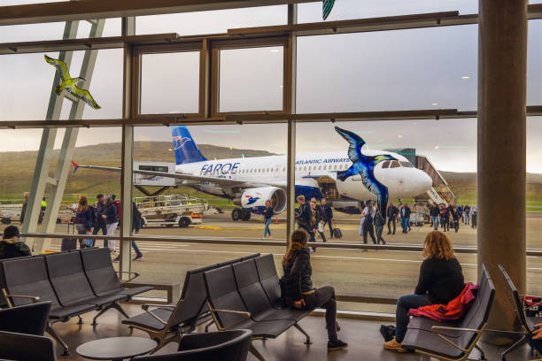 Passengers sit and walk inside an airport terminal while airplanes are parked on the runway outside large windows.