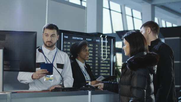 Airport staff assist passengers at a check-in counter, with flight information displayed on screens behind them.