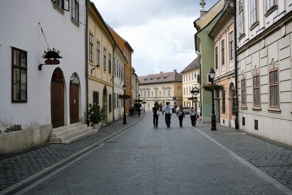 Quiet cobblestone street lined with pastel historic buildings, with a few people walking toward a town square.