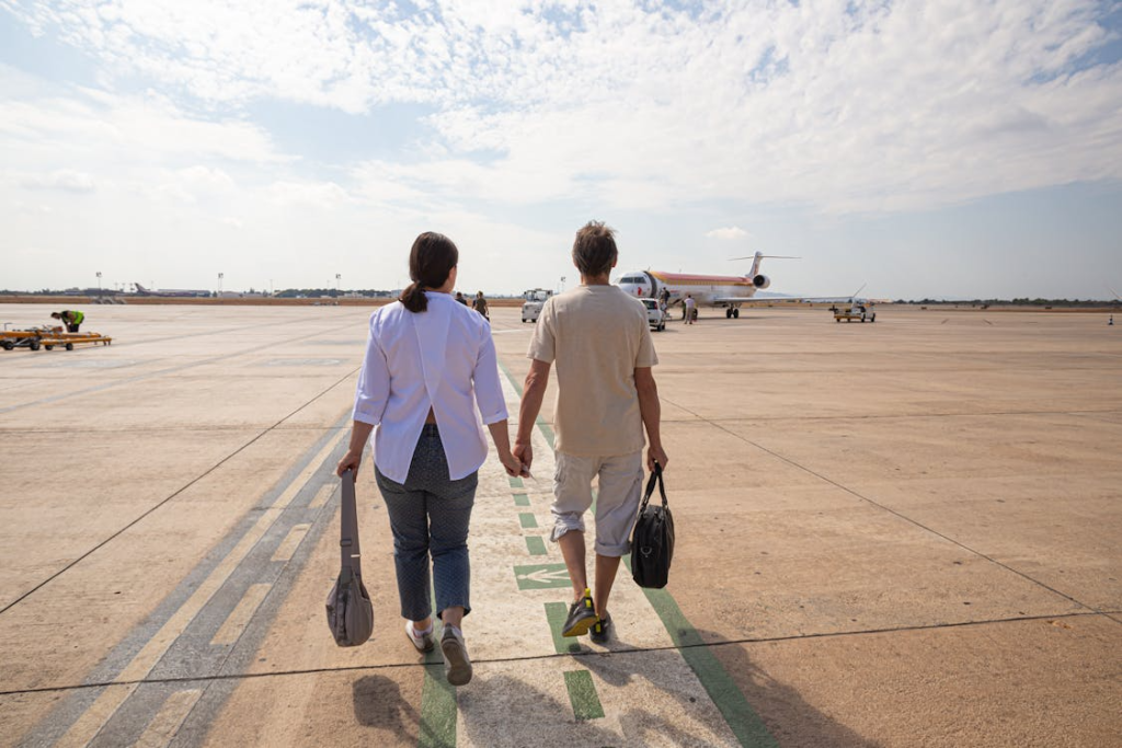 Couple holding hands walks across an airport runway toward a small airplane, carrying travel bags.