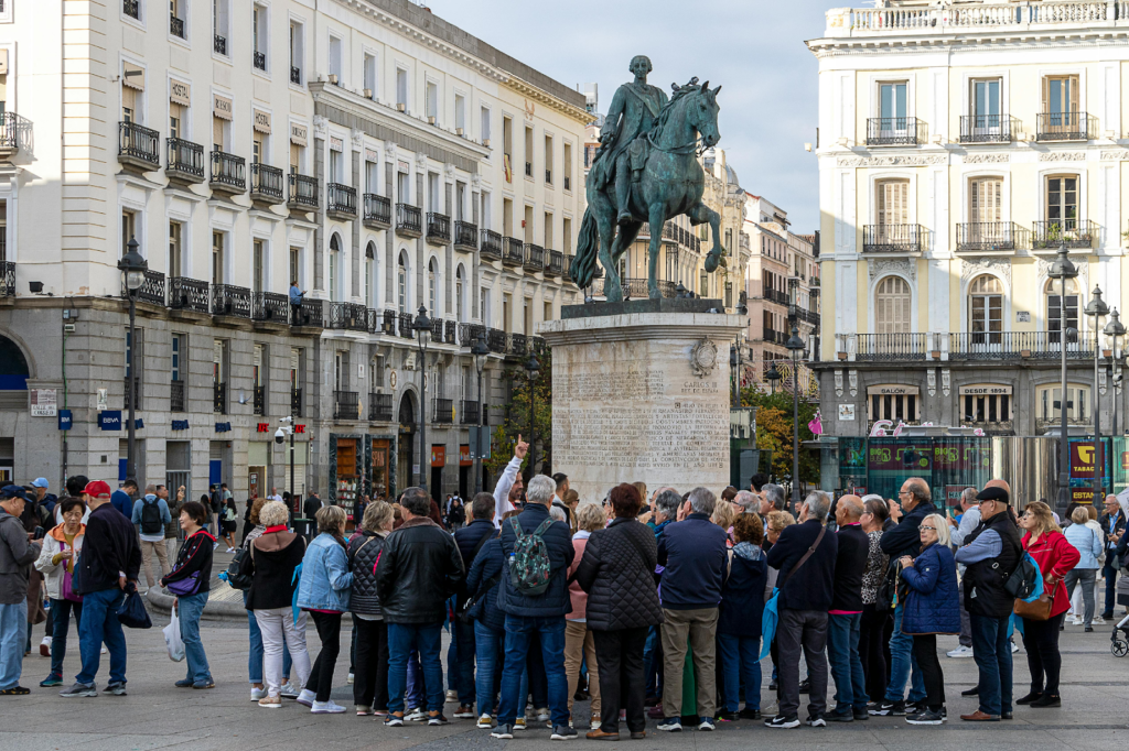 Tourist group gathered around an equestrian statue in a historic city square surrounded by classic European buildings.