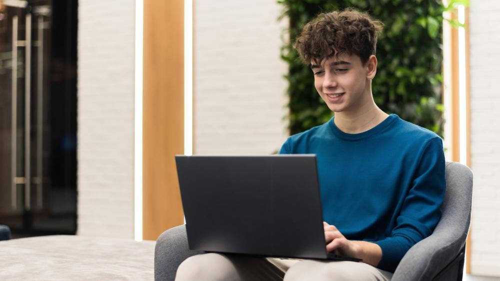 Young person smiling while using a laptop, seated in a chair inside a modern indoor space with plants.
