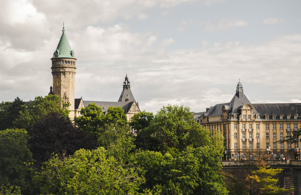 Historic European buildings with a tall stone tower rising above trees under a cloudy sky.