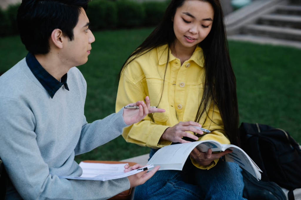 Two college students sitting outdoors, reviewing notes together and discussing a textbook.