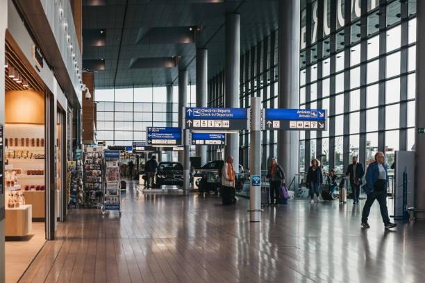 Modern airport terminal interior with travelers walking past shops and overhead directional signs.