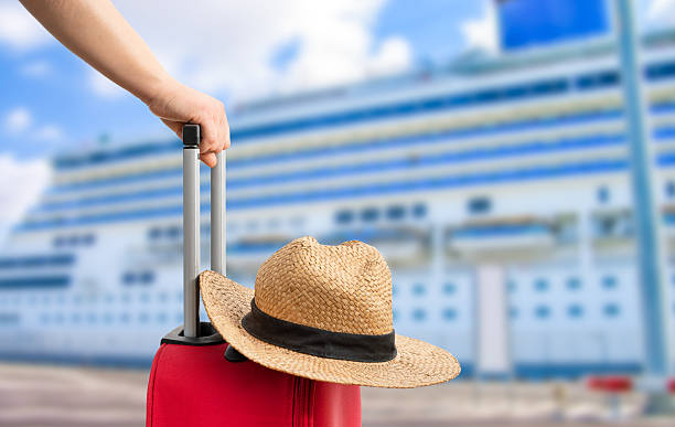 Hand holding a red suitcase with a straw hat, with a large cruise ship docked in the background.