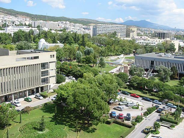 Aerial view of a university campus with modern buildings, green lawns, trees, and parked cars surrounded by a city.