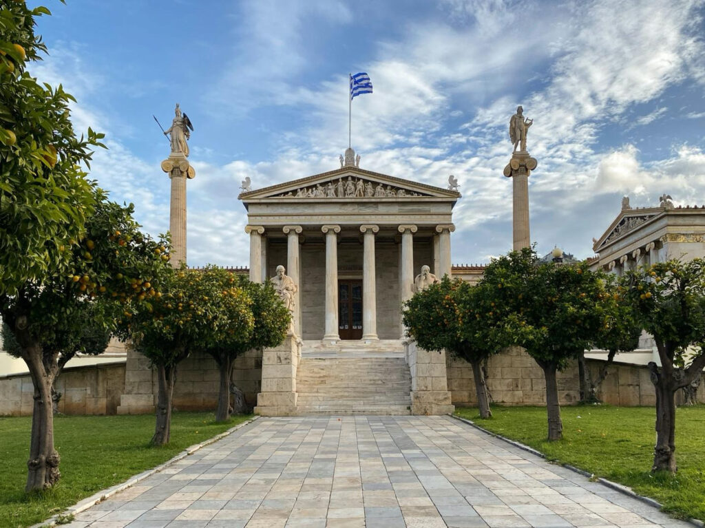 Neoclassical building with tall columns and a Greek flag, framed by statues and orange trees along a stone walkway.