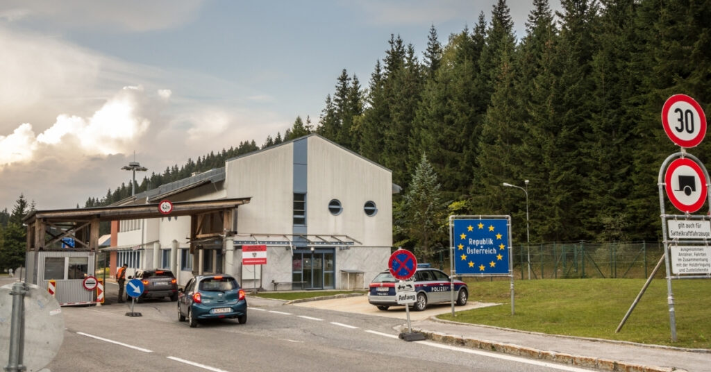 Road border crossing into Austria with EU sign, police car, and vehicles near a forested checkpoint.