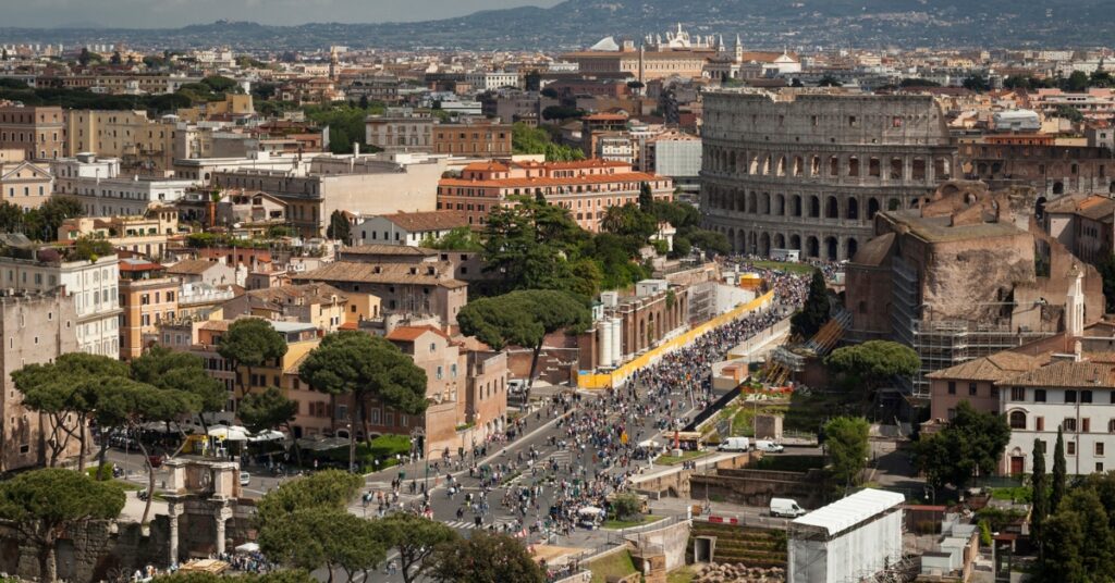 Aerial view of Rome with the Colosseum beside a busy street filled with people and historic buildings.