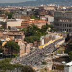 Aerial view of Rome with the Colosseum beside a busy street filled with people and historic buildings.