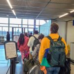 Travelers with backpacks and carry-on luggage waiting in an airport security line near gates.