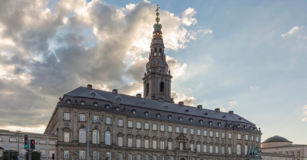 Historic palace with a tall tower in Copenhagen under a cloudy sky, with a city street and passing car in the foreground.