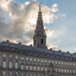 Historic palace with a tall tower in Copenhagen under a cloudy sky, with a city street and passing car in the foreground.