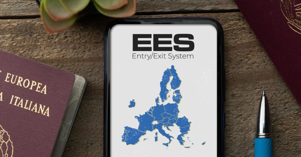 Smartphone screen displays EES Entry/Exit System map of Europe, surrounded by passports and a pen on a wooden surface.