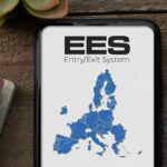 Smartphone screen displays EES Entry/Exit System map of Europe, surrounded by passports and a pen on a wooden surface.