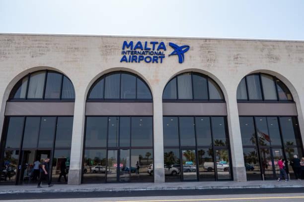 Exterior of Malta International Airport terminal with arched windows, glass entrances, and the airport logo above the facade.