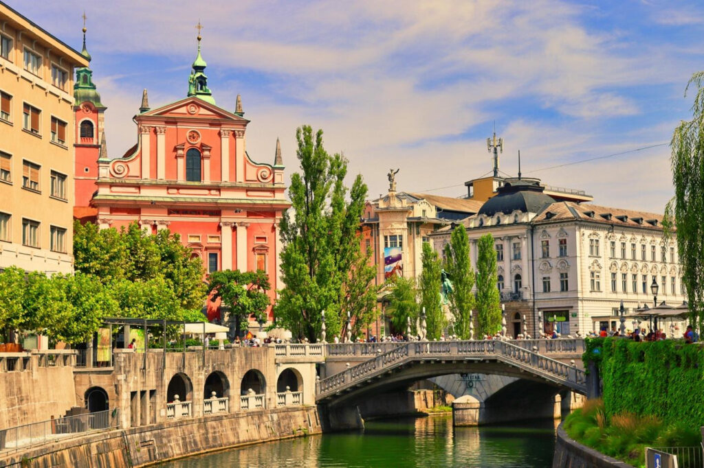 Historic city bridge over a river with colorful buildings and trees along the waterfront.