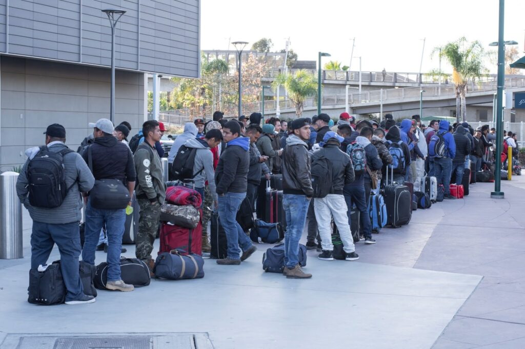 Group of people standing in a long line with suitcases outside a modern building.