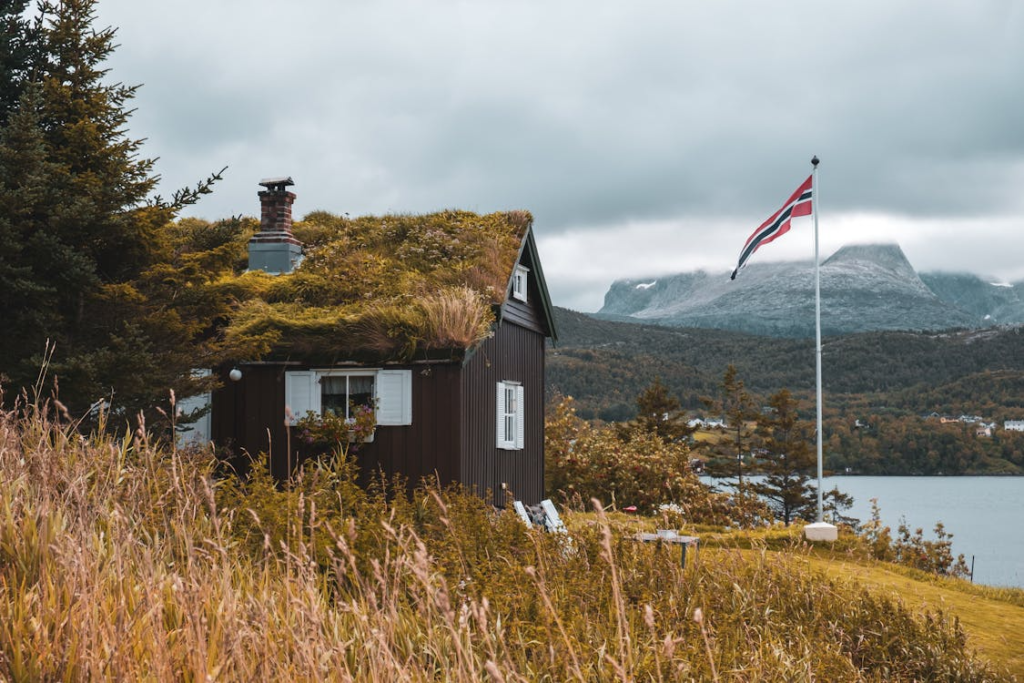 A rustic cabin with a grass-covered roof near a lake, a Norwegian flag on a pole, and mountains in the background.