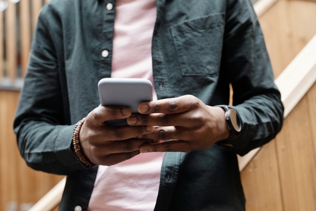 Person holding a smartphone with both hands while standing indoors near a wooden staircase.