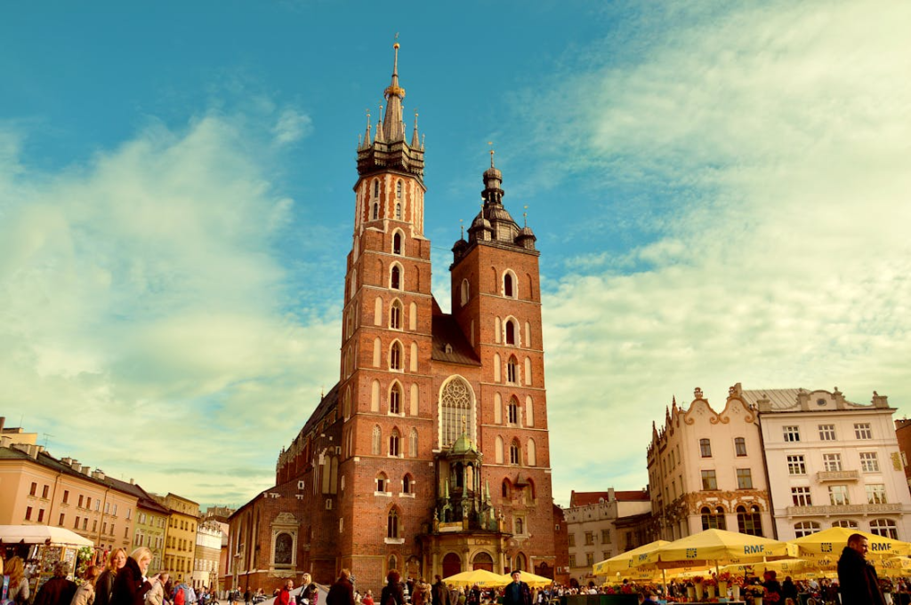 St. Mary’s Basilica with twin towers overlooking Kraków’s Main Square, with people and market umbrellas in the foreground.