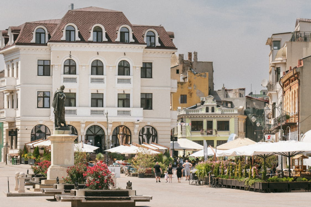 Historic city square with a statue, outdoor cafes, and elegant European buildings surrounding the plaza.