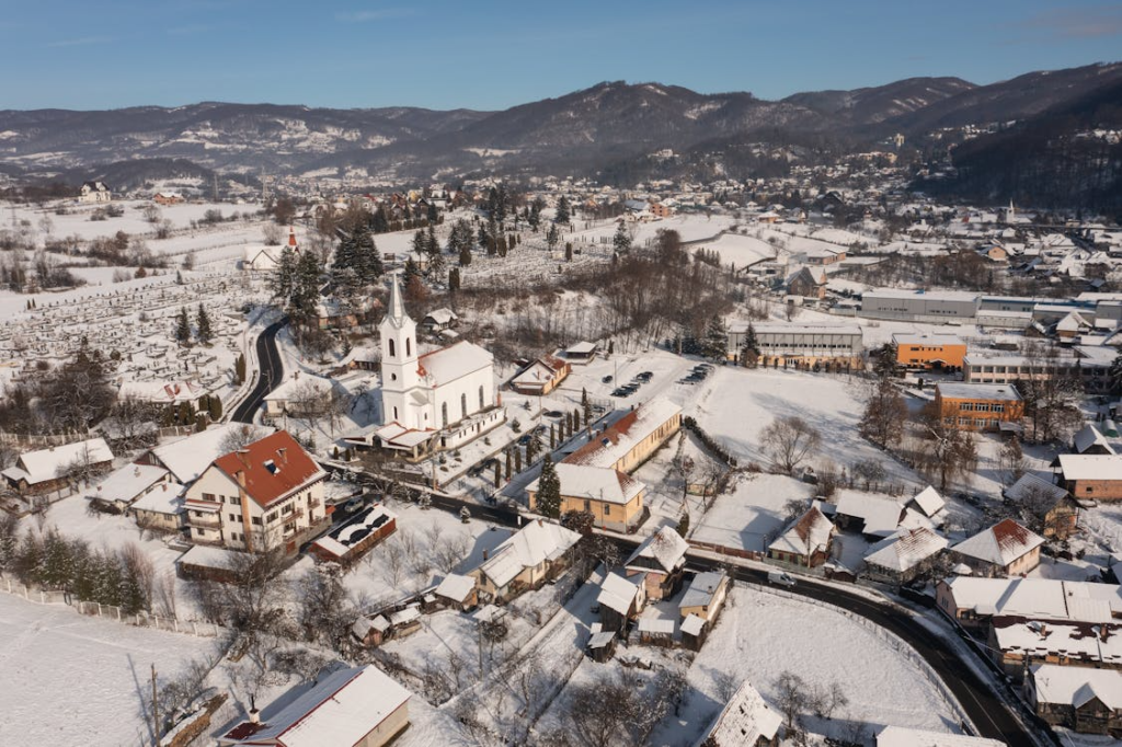 Aerial view of a snowy village with a white church, clustered houses, and mountains in the background.