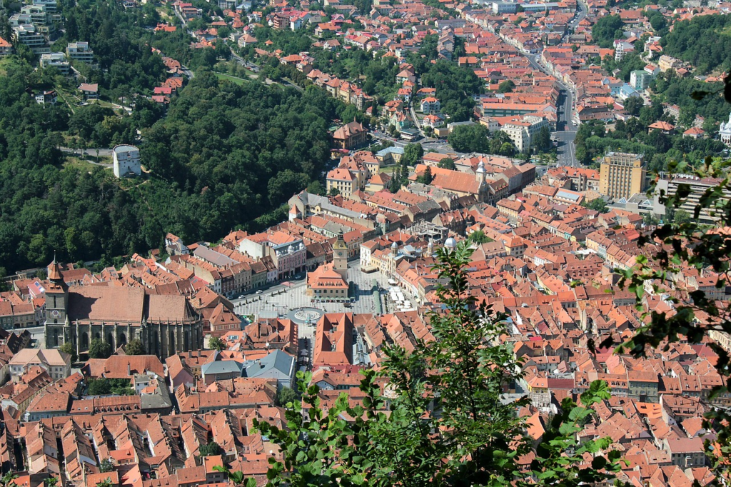 Aerial view of a historic city with red-roofed buildings, a central town square, and surrounding green hills.