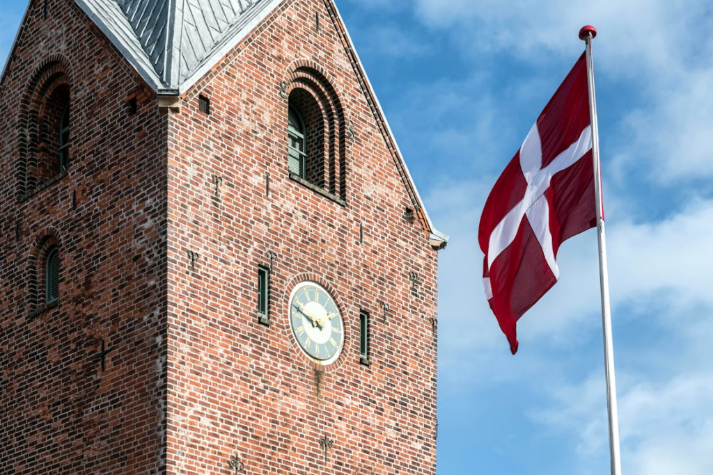 Brick tower with a clock face beside a Danish flag waving against a blue sky.