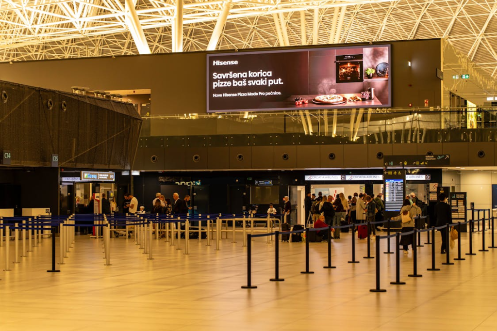 Airport terminal check-in area with passengers waiting in queue lines beneath a large digital advertising screen.
