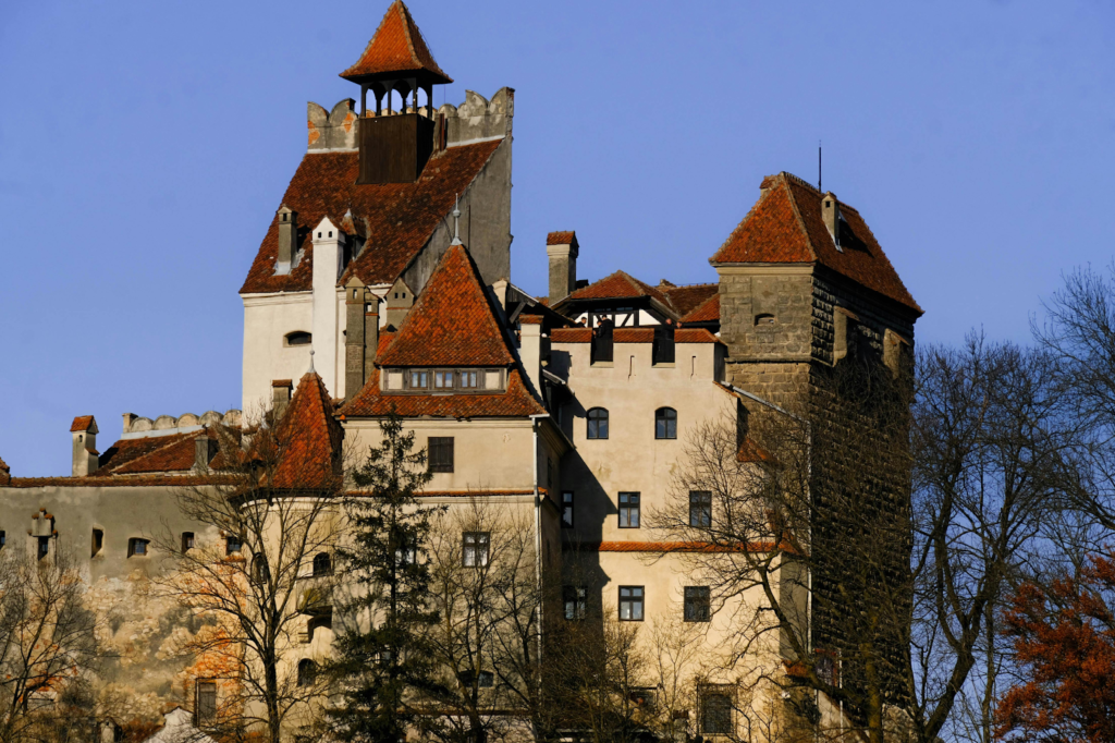 Bran Castle in Romania rises above trees, featuring stone walls and red-tiled roofs against a clear blue sky.