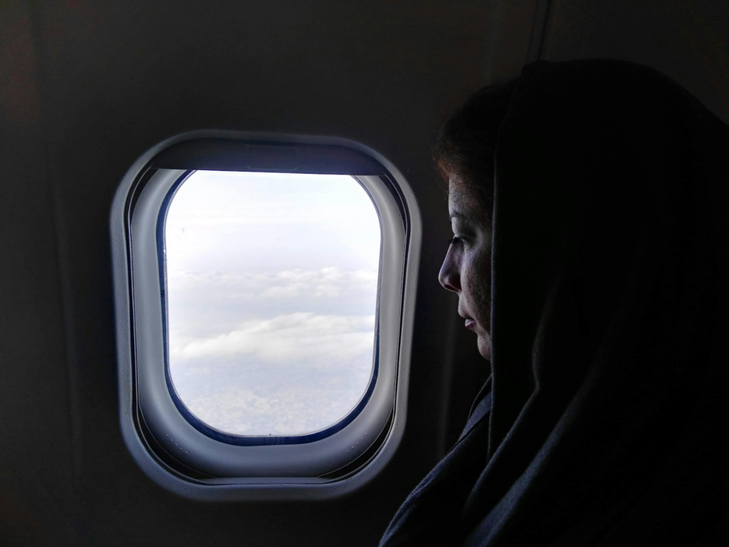 Woman in a headscarf looks out an airplane window at clouds below.