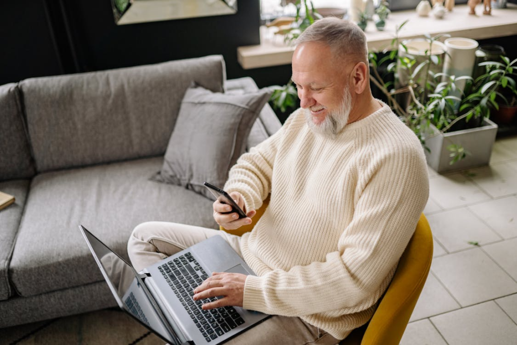 Older man smiling while using a laptop and holding a smartphone, seated in a cozy living room with plants.