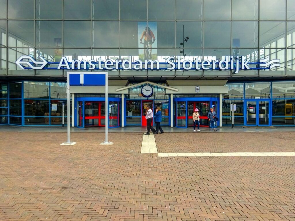 Entrance of Amsterdam Sloterdijk train station with glass facade, blue signage, and people walking outside.